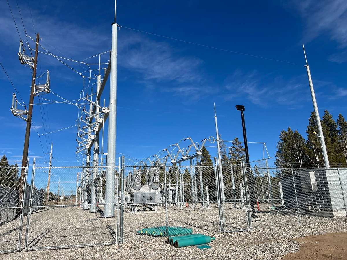 An existing fenced electrical substation with steel switch structures and power lines under a blue sky