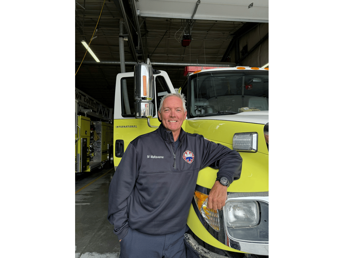 Firefighter in a jacket embroidered "M Maltaverne" leaning against a yellow-green fire engine in a station bay