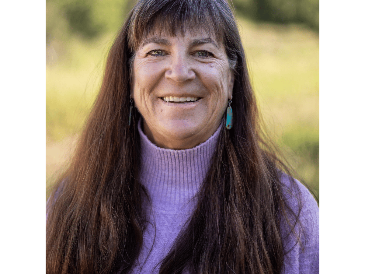 Sue Muncaster smiling outdoors in a lavender sweater and turquoise earrings