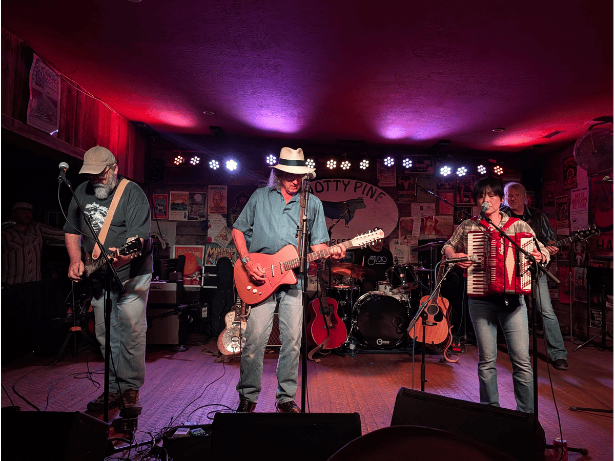 James McMurtry and his bandmates performing on the Knotty Pine stage under red lights, with guitars and an accordion