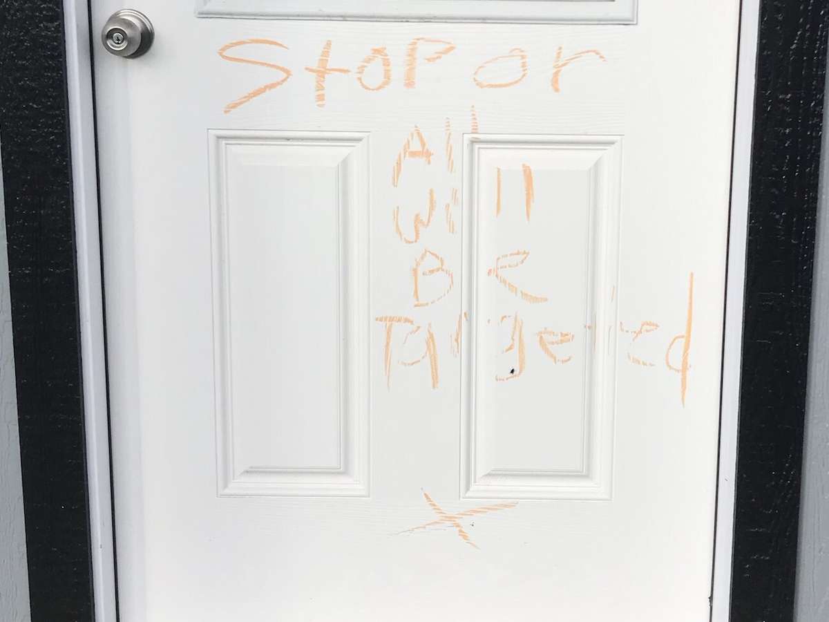The words "Stop or All Will Be Targeted" written in orange on the white door of a small building under construction, with an X below.