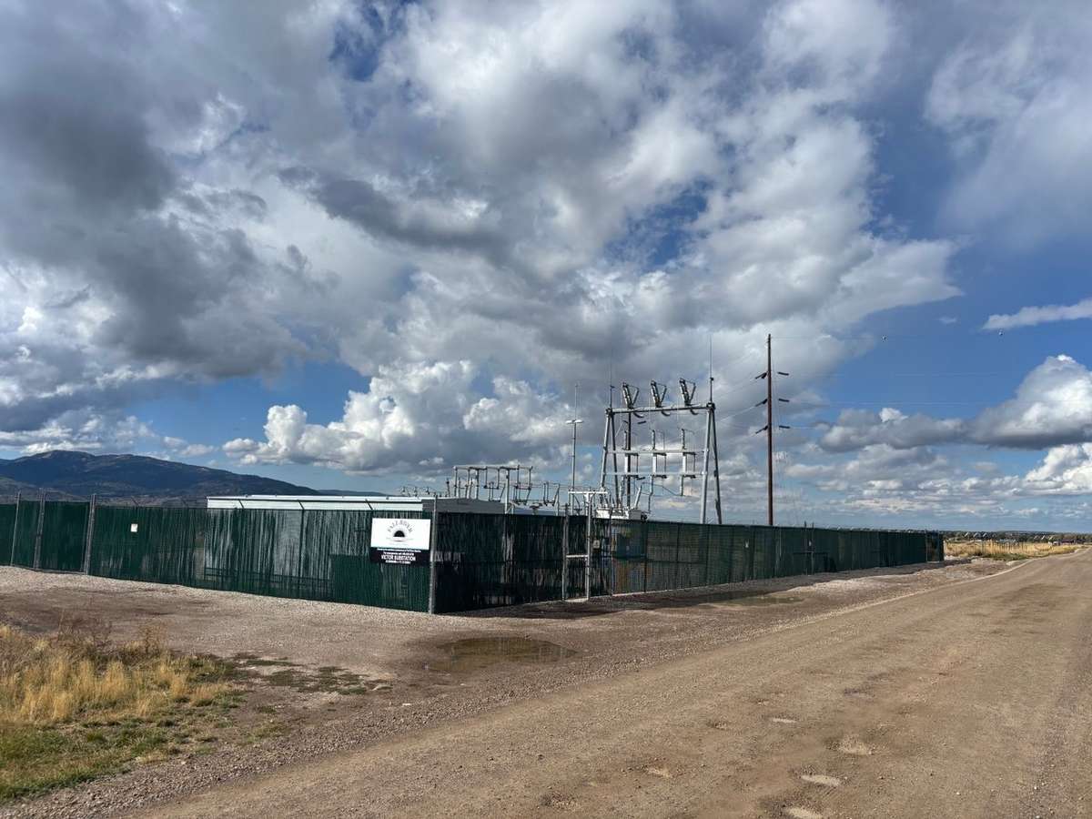An existing small fenced electrical substation beside a gravel road under a cloudy sky, with hills in the distance